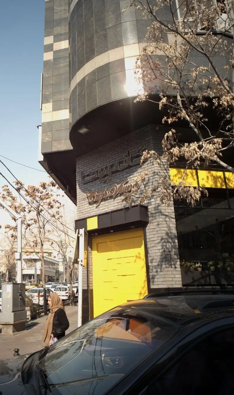 نمای بیرونی کافه ویونا، تابلوی Cafe Viyuna روی دیوار آجری، درب زرد برجسته، ساختمان مدرن خاکستری و زن رهگذر در خیابان.