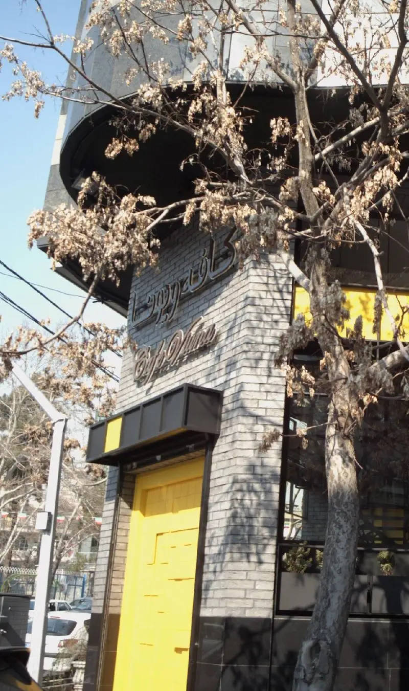 نمای بیرونی کافه ویونا در تهران با درب ورودی زرد رنگ، طراحی مدرن و آجر خاکستری. یک درخت با برگ‌های خشک در پیش‌زمینه.