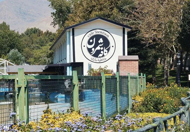 کافه شمرون تهران، ساختمانی با تابلوی "Cafe Shemroon" و "خاطره بازی" در محیطی سرسبز، پارک مانند و کوهستانی.