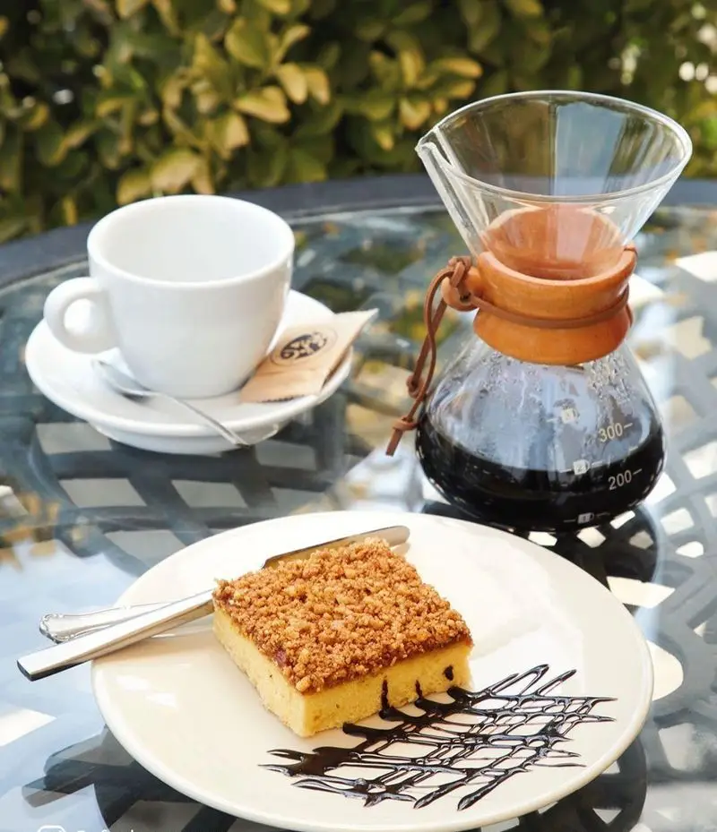 یک میز دلنشین در کافه شمرون: قهوه دمی تازه در کمکس، کیک کرمبل خوشمزه و فنجان خالی آماده لذت بردن در فضای باز.