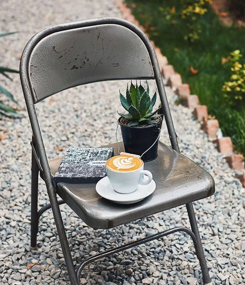 کافه شمرون: فنجان لاته آرت با طرح هنری، کتاب تهران و گلدان کوچک روی صندلی فلزی در فضای باز. محیطی دلنشین برای آرامش.