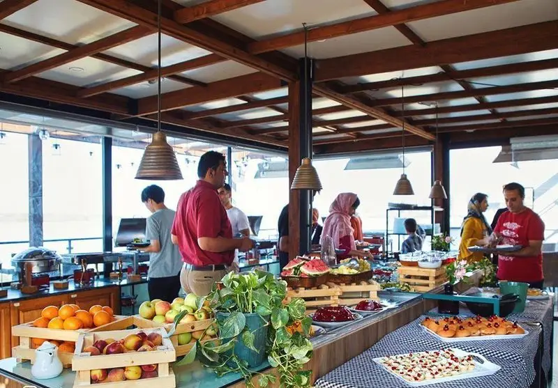 مشتریان در حال لذت بردن از بوفه صبحانه و ناهار کافه شمرون با میوه‌های تازه، دسر و غذاهای باکیفیت در محیطی دلنشین و دکوراسیون چوبی زیبا.