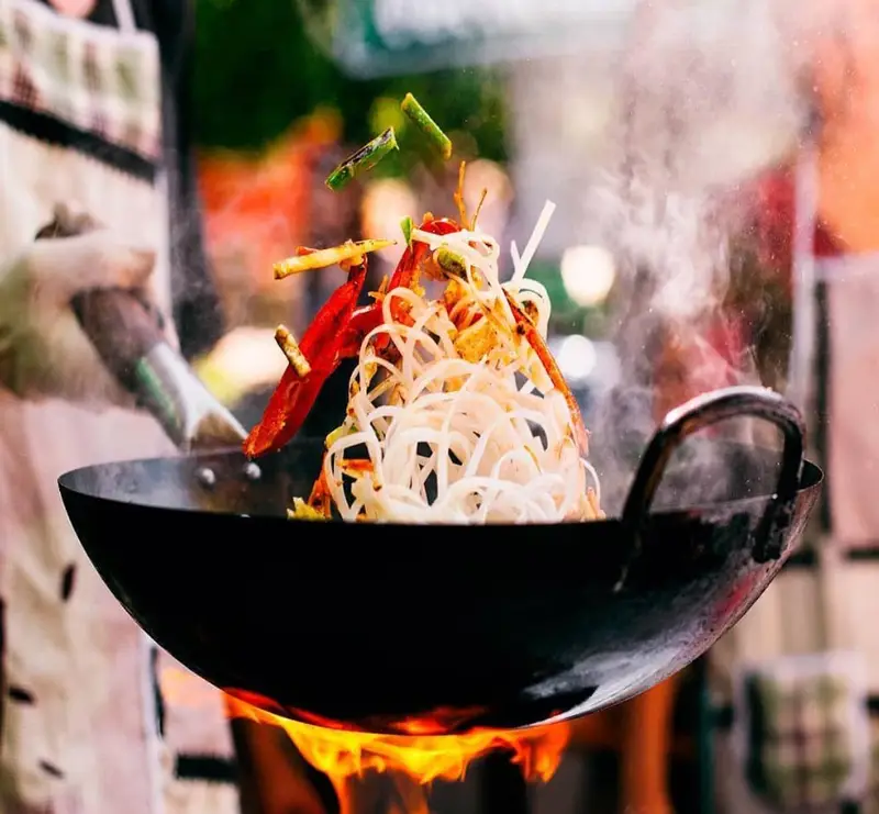 ووک‌گردانی ماهرانه نودل و سبزیجات رنگارنگ در شعله‌های آتش، با بخار داغ، هنر آشپزی پرشور پیتزا سرخاب سیب.