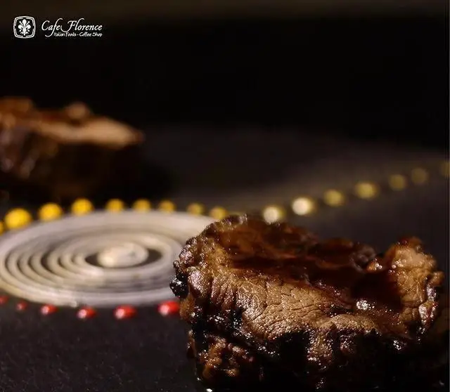 کافه فلورانس: تصویری نزدیک از تکه گوشت لذیذ و آبدار، تزئین شده با سس براق و چیدمان هنری روی بشقاب مشکی. غذای اصلی و شیک.