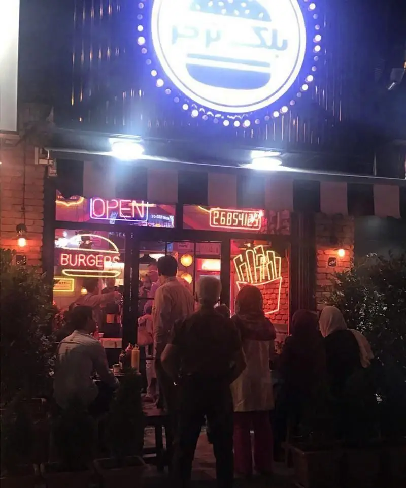 نمایی از رستوران بلک برگر در شب، با تابلوهای نئون روشن "OPEN" و "BURGERS"، لوگوی برجسته و مشتریانی در فضای بیرونی.