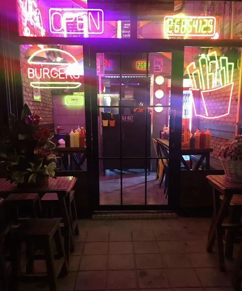ورودی شبانه رستوران بلک برگر با نورپردازی نئون بنفش "OPEN"، تابلوهای نئون برگر و سیب زمینی سبز، میز و صندلی‌های چوبی.