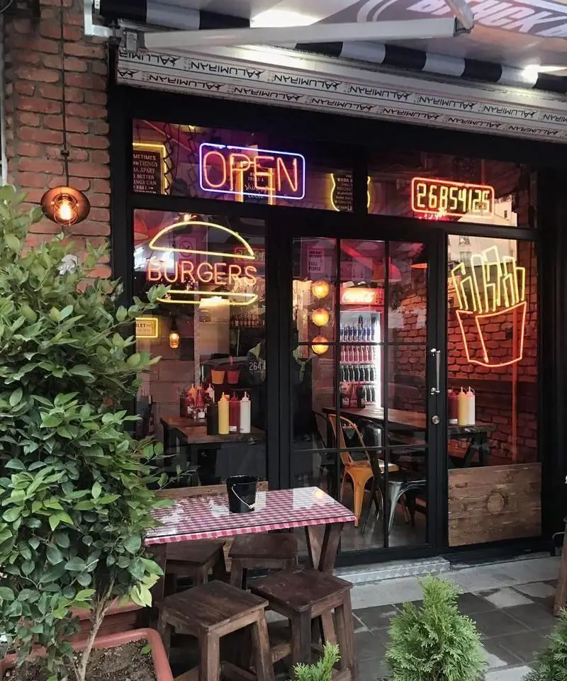 نمای بیرونی رستوران فست فود بلک برگر با تابلوهای نئونی "OPEN"، "BURGERS" و سیب‌زمینی، میزهای چوبی و فضای نشیمن جذاب.