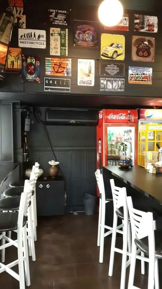فضای داخلی کافه برگر ن با دکوراسیون دیواری پر از تابلوهای وینتیج، میز پیشخوان با صندلی‌های سفید و یخچال نوشیدنی.