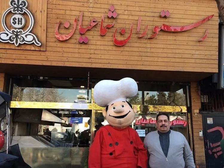 رستوران شماعیان: تابلوی چوبی با نام فارسی و لوگوی SH، سرآشپز کارتونی و صاحب رستوران آماده پذیرایی از مشتریان با غذای ایرانی.