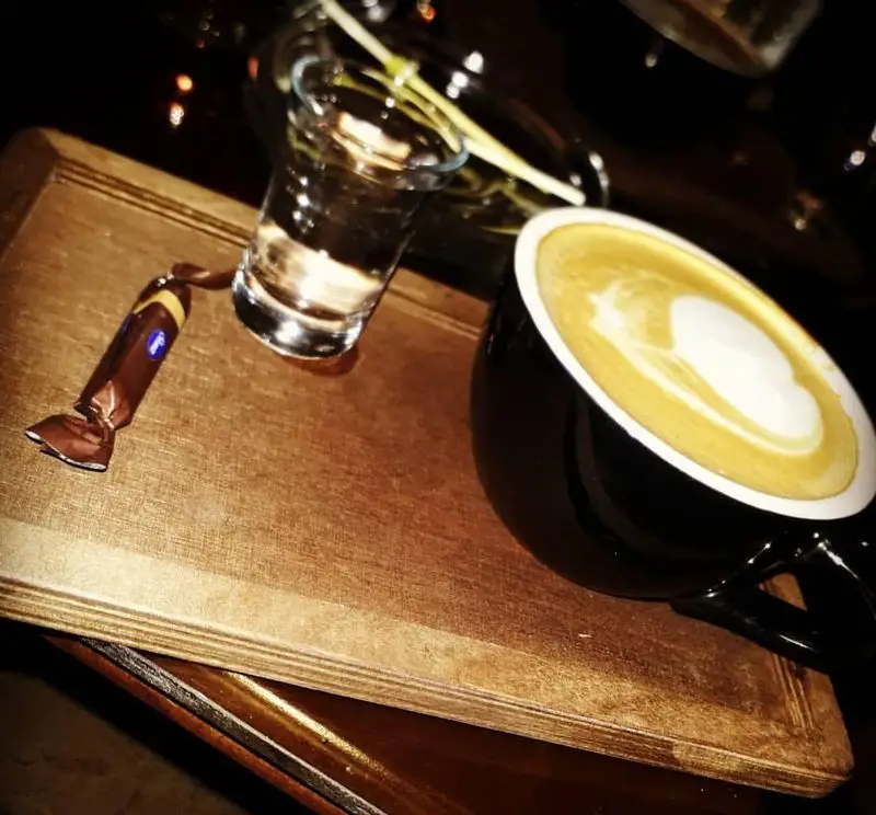 قهوه لاته آرت زیبا، یک لیوان آب و آبنبات روی سینی چوبی در محیط دلنشین کافه دایموند. لحظاتی آرام با نوشیدنی دلچسب در کافه دایموند.