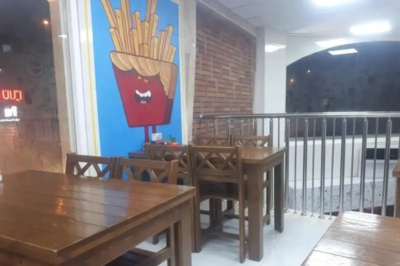 فضای داخلی دلنشین فست فود ممدخان، شامل میز و صندلی چوبی، دیوار طرح آجر و نقاشی دیواری خلاقانه کاراکتر سیب زمینی سرخ کرده خندان.