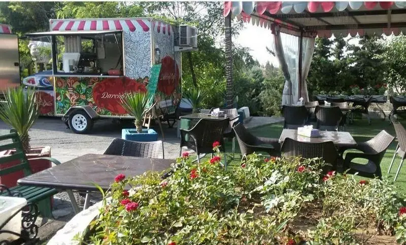 کافه سیار دارچینی در فضای باز سرسبز با میز و صندلی حصیری، آماده سرو انواع نوشیدنی و خوراکی در محیطی دلنشین.