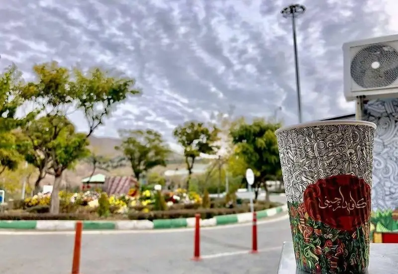 عکس یک لیوان قهوه طرح دار از کافه دارچینی در فضای باز پارک با درختان سرسبز، گل‌ها و آسمان ابری، محیطی دلنشین برای نوشیدنی.