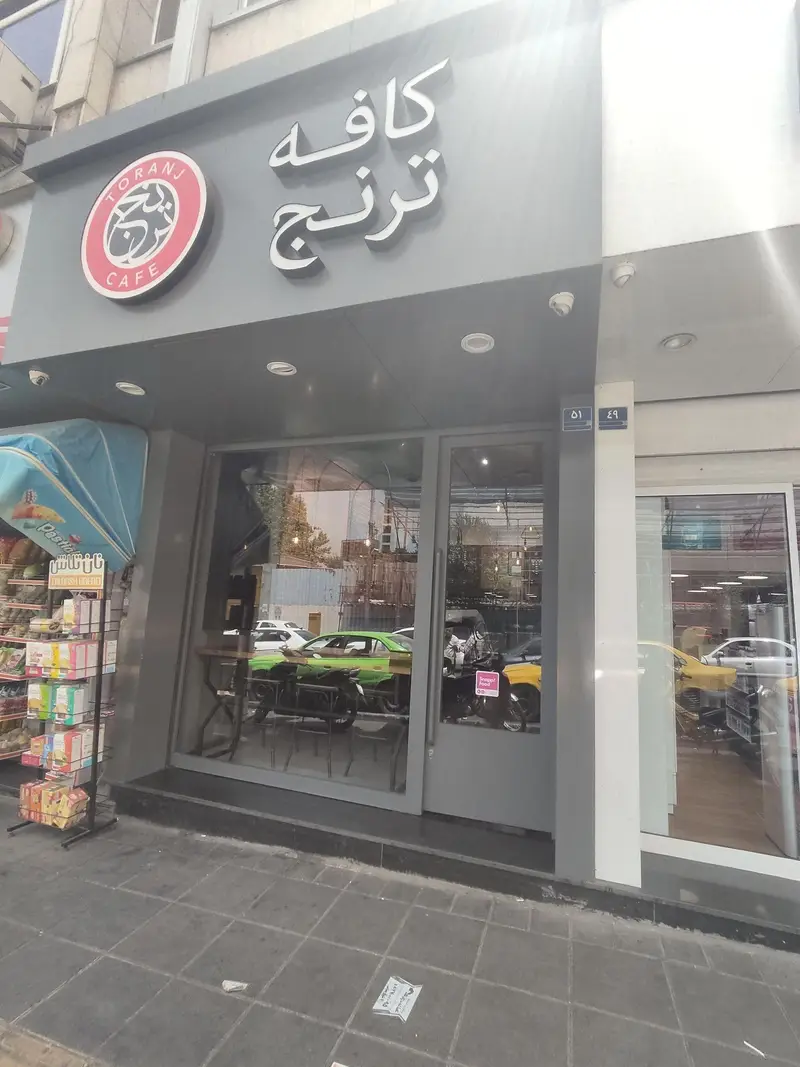 عکس نمای خارجی کافه ترنج (Toranj Cafe) در روز، با تابلو لوگوی برند و ورودی شیشه‌ای. فضایی دنج برای قهوه و دورهمی در تهران.