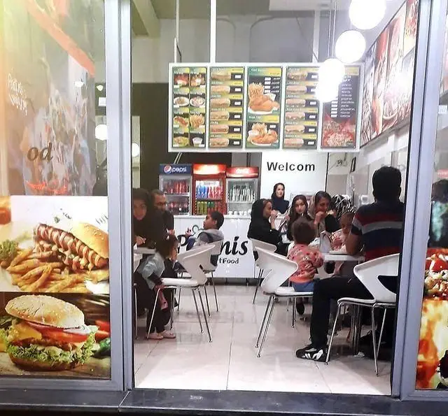 فست فود پودینیس، نمای داخلی شلوغ با خانواده‌ها و مشتریانی که از منوی متنوع شامل برگر، هات‌داگ و مرغ سوخاری لذت می‌برند.