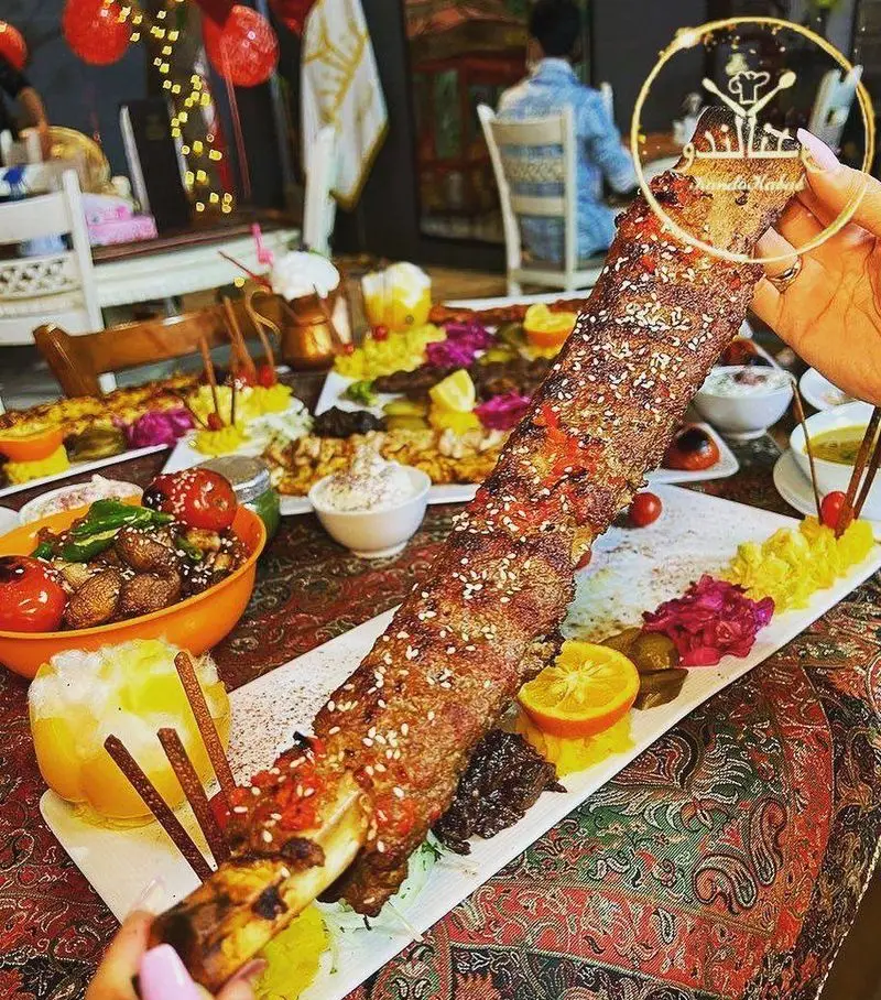 کندو کباب: دنده کباب بلند و استخوان‌دار با چاشنی کنجد، در کنار انواع غذاها و دورچین‌های رنگارنگ در رستورانی سنتی.