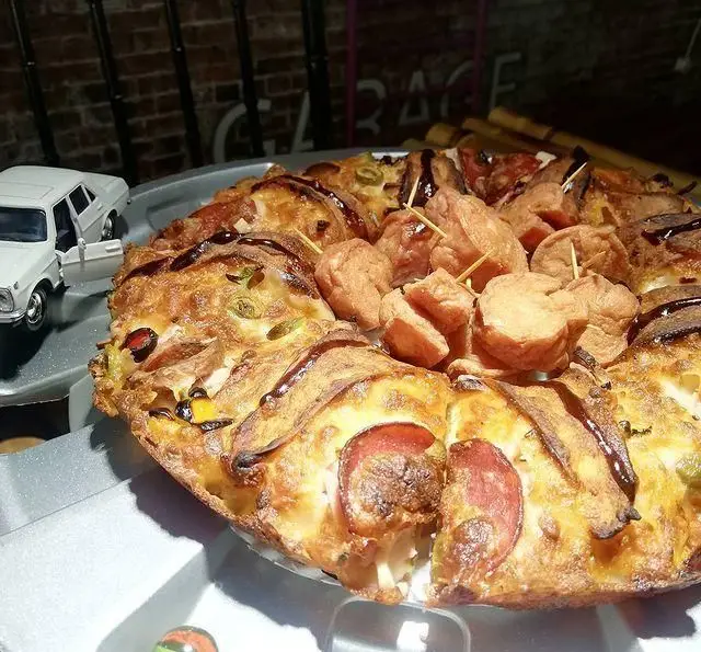 تصویری از پیتزای ویژه و خلاقانه فست فود گاراژ، شامل برش‌های سوسیس و کالباس، زیتون و سس قهوه‌ای. غذایی خاص و خوش طعم برای شما.