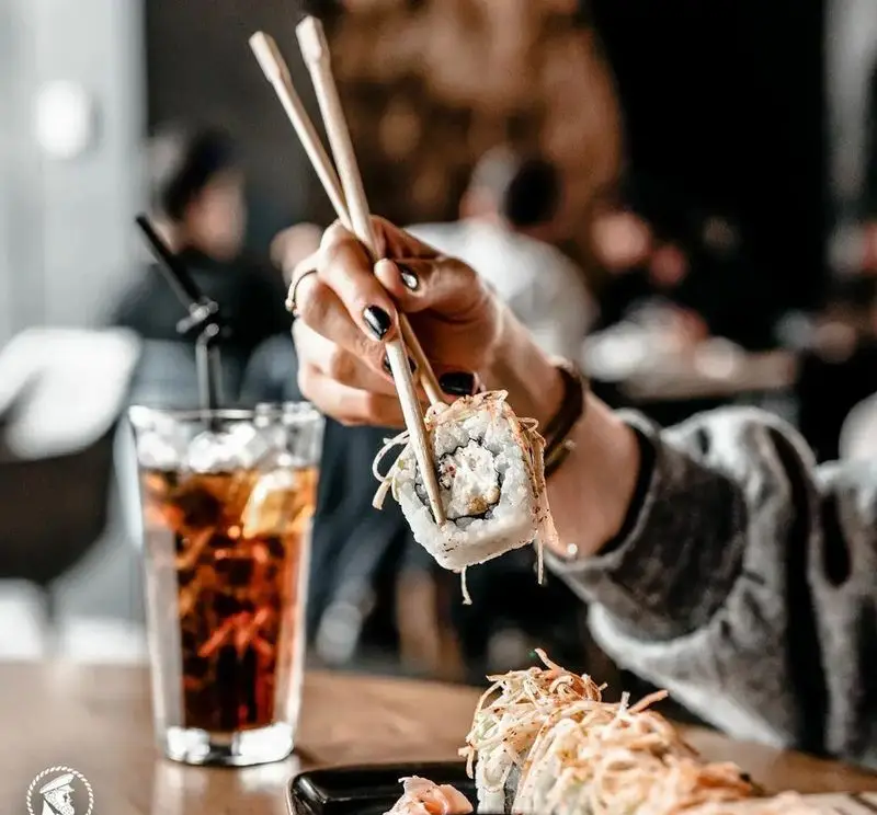شخصی در کافی شاپ کاپیتان لانژ در حال خوردن رول سوشی با چاپستیک، همراه با یک نوشیدنی خنک. تجربه غذایی دلپذیر در کاپیتان لانژ.
