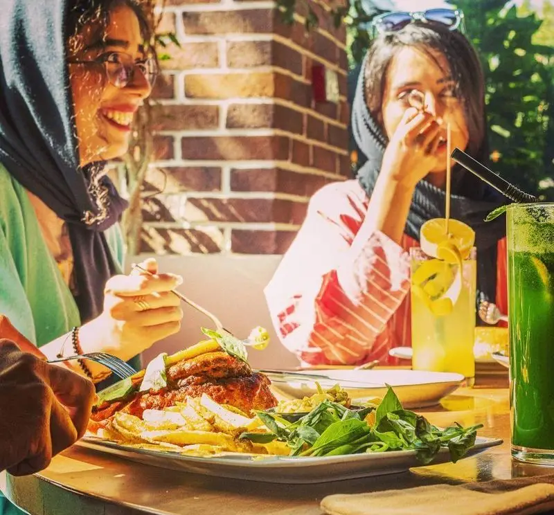 دو خانم با حجاب در کافی شاپ کاپیتان لانژ در حال صرف غذا (شامل سیب زمینی و سالاد) و نوشیدنی‌های خنک در فضایی دلنشین و آفتابی.