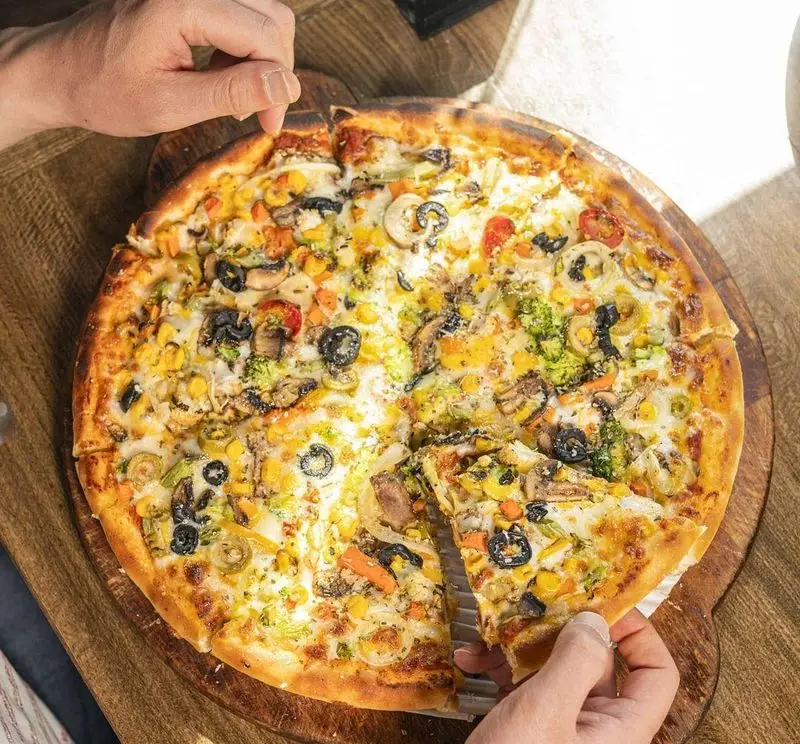 پیتزا سبزیجات دست‌ساز و تازه کاپیتان لانژ با ذرت، قارچ، زیتون، بروکلی و هویج. سرو شده روی میز چوبی در کافی شاپ.