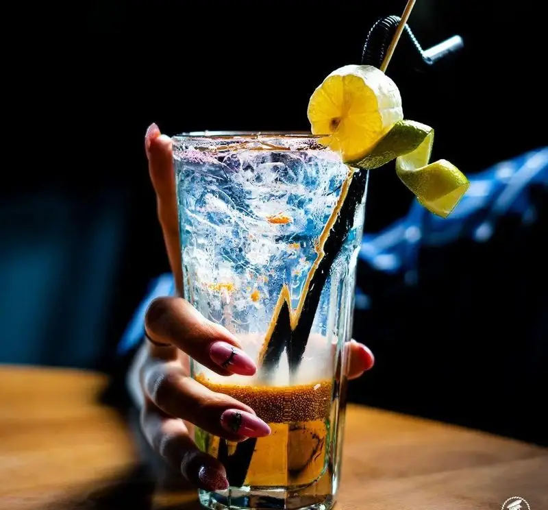 دست خانمی با ناخن‌های صورتی، نوشیدنی خنک و لایه‌دار کافی شاپ کاپیتان لانژ را با یخ، لیمو و تزیینات زیبا در لیوان بلند گرفته است.