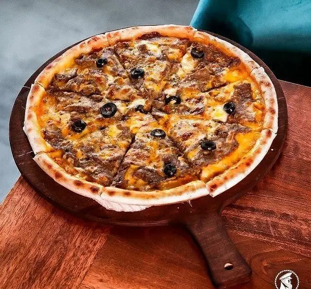 پیتزای آمریکایی گوشت و زیتون سیاه تازه کاپیتان لانژ، با پخت عالی و نان ترد، سرو شده روی تخته چوبی شیک در کافی شاپ.