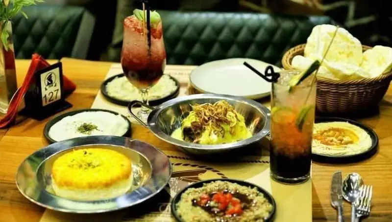 میز پر از غذاهای متنوع و نوشیدنی‌های تازه در کافی شاپ کاپیتان لانژ، شامل حمص، دیس‌های لذیذ و کوکتل‌های خوش‌رنگ و شیک.