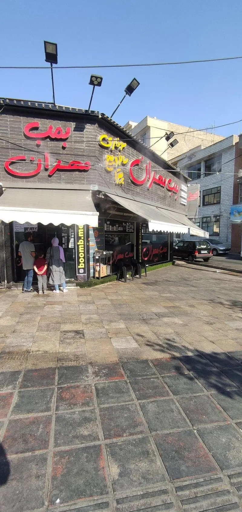 فست فود بمب مهران، یک رستوران برگر و ساندویچ پرطرفدار با تابلوی جذاب، مشتریان منتظر و محوطه بیرونی برای پذیرایی در طول روز.