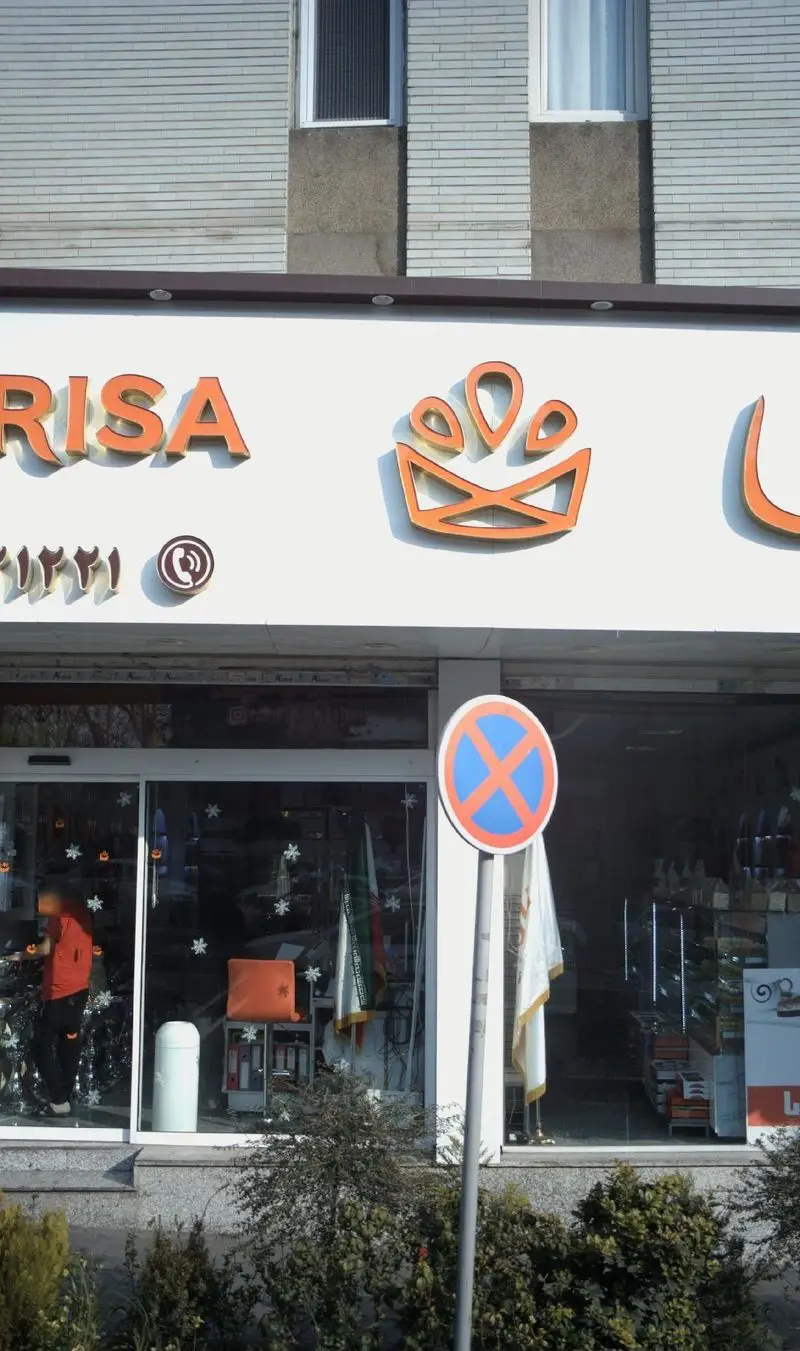 نمای بیرونی فروشگاه شیرینی لاریسا با تابلوی برجسته نارنجی، لوگوی خاص و شماره تماس، به همراه ویترین پر از شیرینی و دسر.