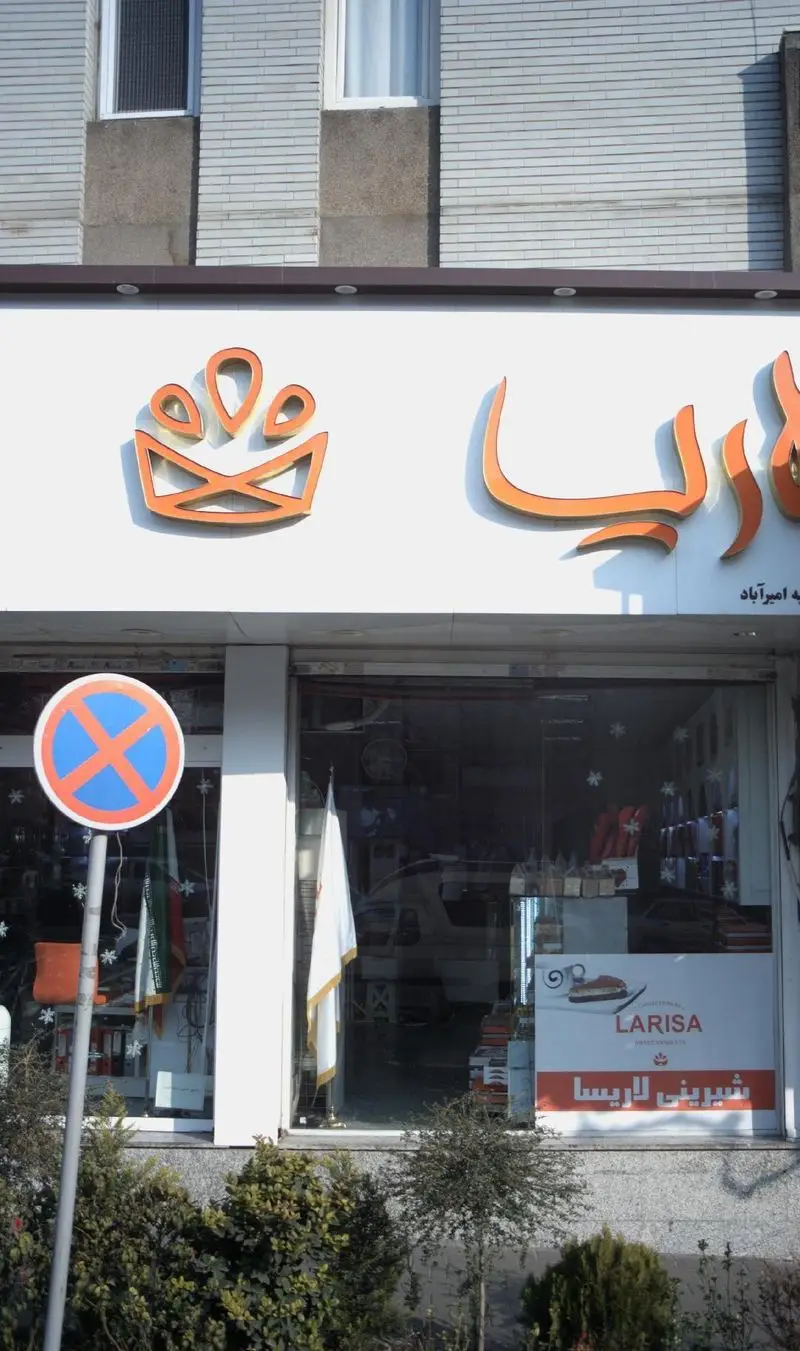 نمای بیرونی و تابلو اصلی شیرینی لاریسا با آرم تاج نارنجی و بنر تبلیغاتی دسرها در ویترین، یک قنادی مدرن و شیک.