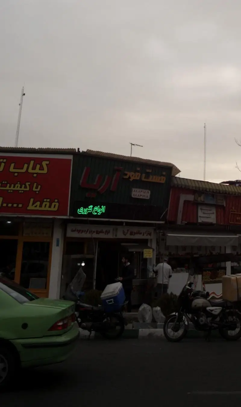 تصویر نمای بیرونی فست فود آریا با تابلوهای کبابی و انواع کباب، شامل موتورسیکلت‌های پیک و مشتریان در جلوی رستوران.