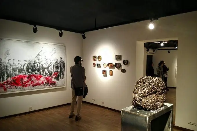 گالری هنری آ: یک بازدیدکننده در حال تماشای نقاشی بزرگ با رنگ قرمز و مجموعه ای از آثار دیواری و یک مجسمه خاص در فضای نمایشگاه.