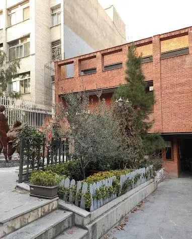 گالری آ: نمای بیرونی ساختمان آجری با پنجره های بلند، باغچه زیبا، درختان سبز و نرده های تزئینی. فضایی هنری و دلپذیر.