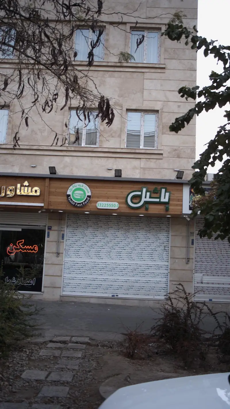 تصویری از نمای ساختمان فست فود یاشیل با تابلوی سبز رنگ، لوگوی برگر و شماره تماس ۳۳۲۲۵۵۵۰-۱. درب بسته فروشگاه در یک روز ابری.