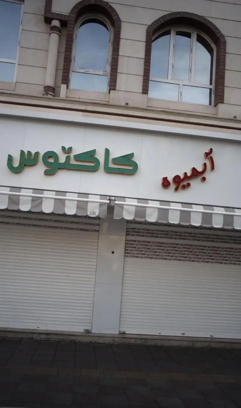 عکس نمای مغازه آبمیوه، بستنی کاکتوس با تابلوهای سبز و قرمز، کرکره های بسته و آلاچیق راه راه.