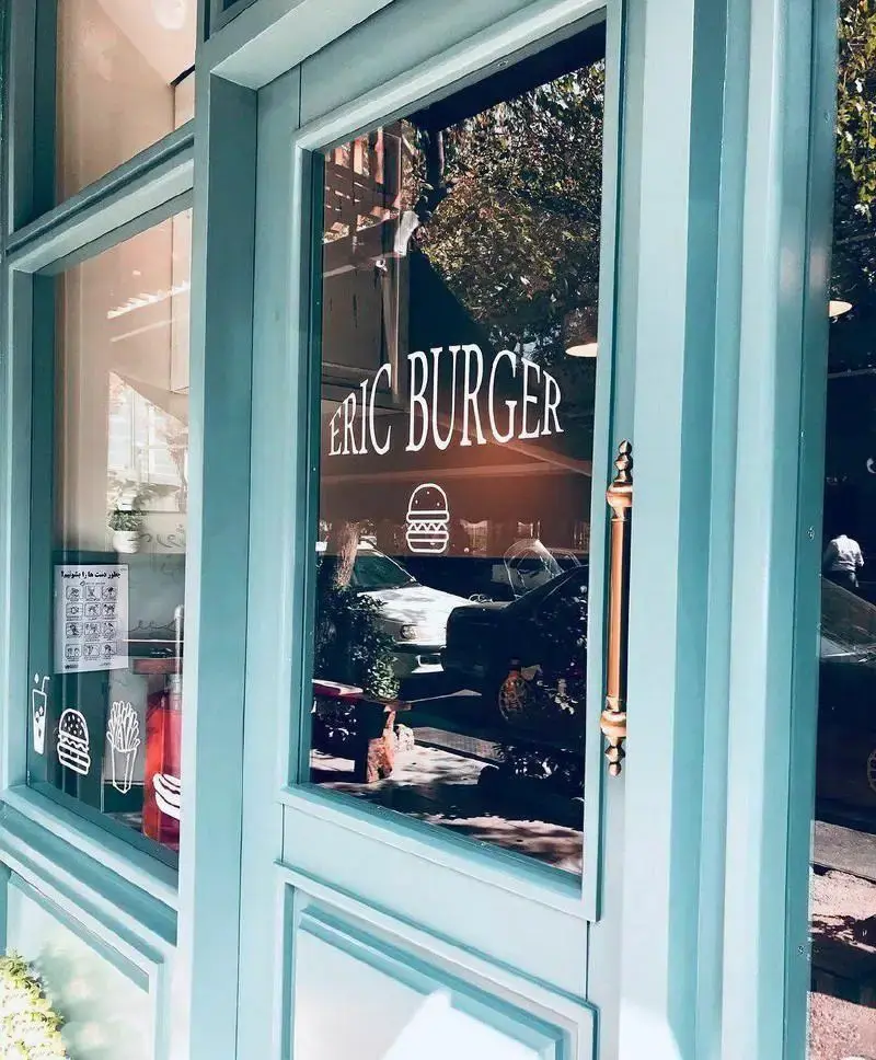 نمای خارجی رستوران فست فود اریک برگر با پنجره‌ها و درب فیروزه‌ای، لوگوی Eric Burger و آیکون‌های برگر، سیب زمینی و نوشیدنی روی شیشه.