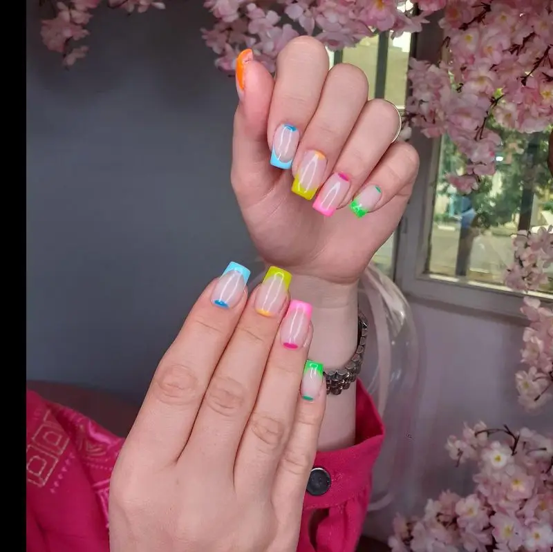 تصویر طراحی ناخن فرنش چندرنگ و رنگارنگ روی دست مشتری با پسزمینه شکوفههای صورتی در سالن زیبایی ساناز صفری