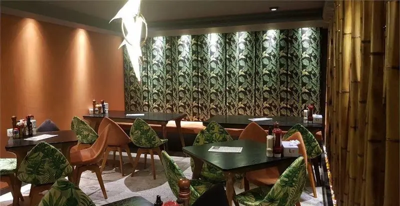 فضای داخلی دلنشین رستوران میخوش با چیدمان میز و صندلی‌های شیک، پرده‌های طرح‌دار برگ سبز و دیوار دکوراتیو بامبو، ایده‌آل برای یک وعده غذایی خاص.