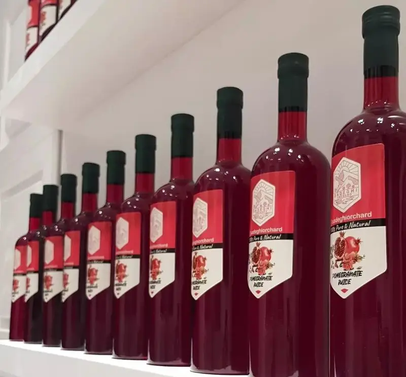 ردیفی از بطری‌های آب انار ۱۰۰٪ طبیعی و خالص باغ صادقی مقدم بر روی قفسه، محصولی مرغوب از کسب و کار انار و آب انار صادقی مقدم.