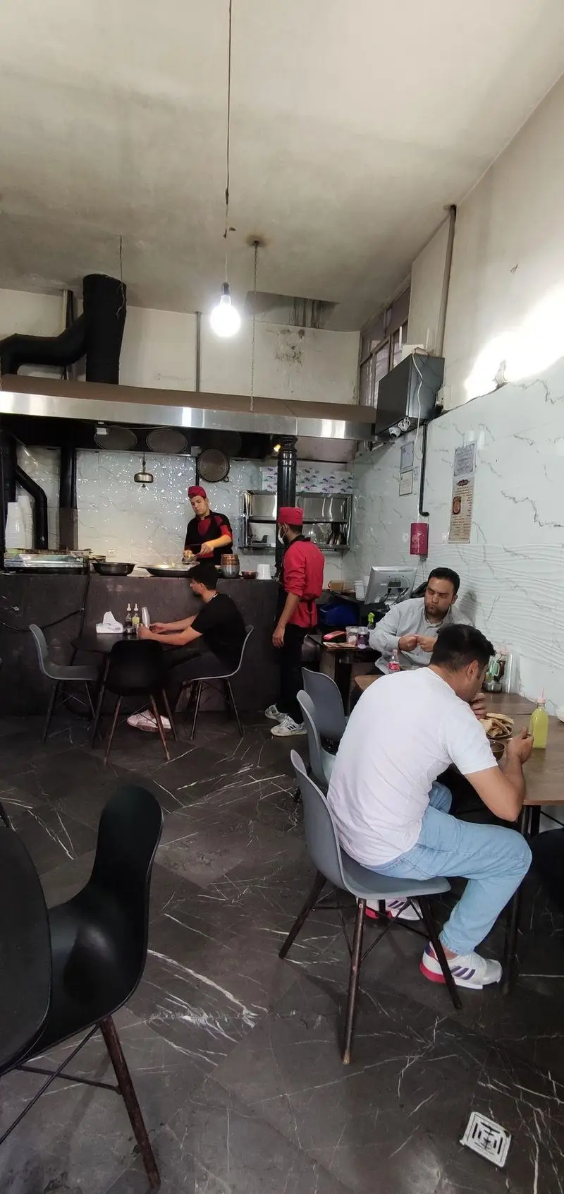 تصویر داخلی طباخی توکلی: آشپزانی ماهر در آشپزخانه باز مشغول آماده‌سازی غذا و مشتریان در فضایی دلنشین مشغول صرف وعده غذایی.