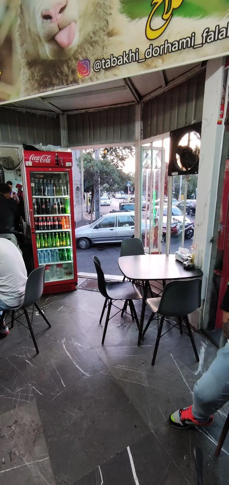 فضای داخلی طباخی توکلی، با یخچال کوکاکولا پر از نوشیدنی، میز و صندلی برای مشتریان. تابلوی بالای سر با تصویر گوسفند و پیج اینستاگرام. منظره خیابان از پنجره.