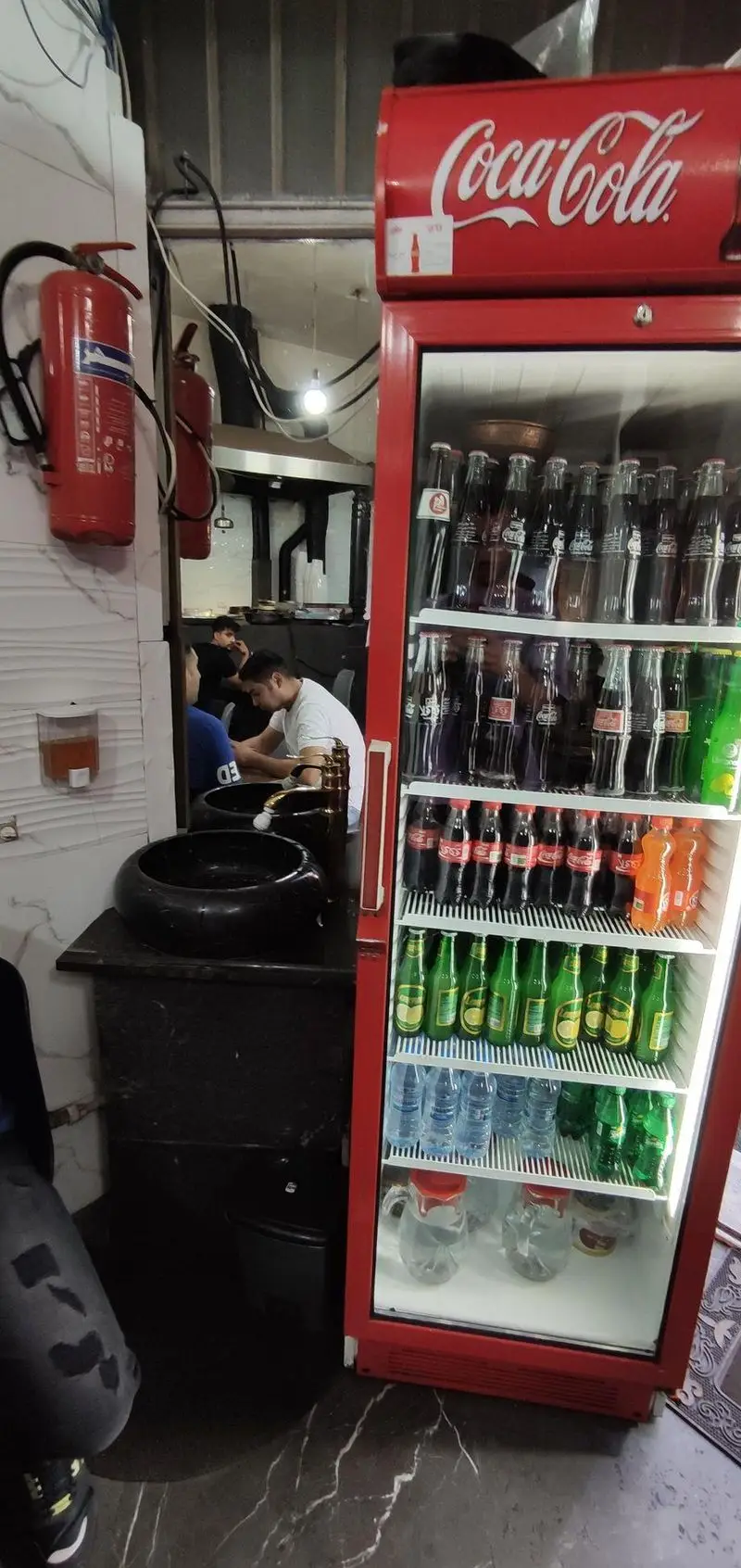 فضای داخلی طباخی توکلی، با یخچال بزرگ کوکاکولا پر از نوشیدنی‌ها، کپسول آتش‌نشانی و سینک شستشو. مکانی مناسب برای پذیرایی.