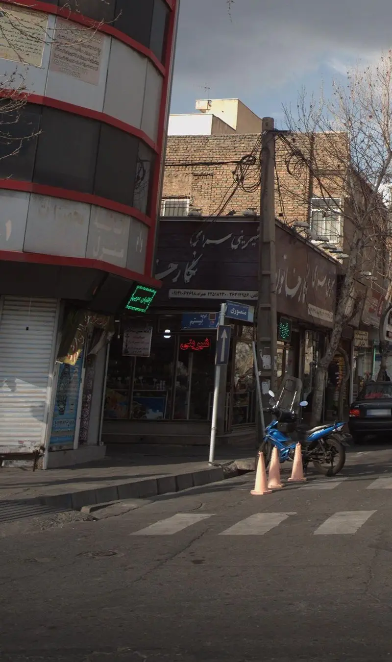 تصویر نمای بیرونی شیرینی سرای نگارخاتون در کنار سایر کسب و کارها، به همراه موتورسیکلت و علائم ترافیکی در خیابان منصوری.