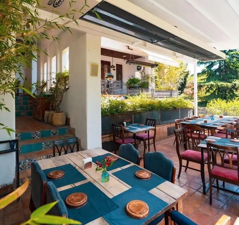 تراس دلپذیر رستوران مس توران با میزهای چوبی زیبا، رومیزی‌های آبی، گلدان‌های گل، صندلی‌های راحت و فضایی سرسبز و آرامش‌بخش.