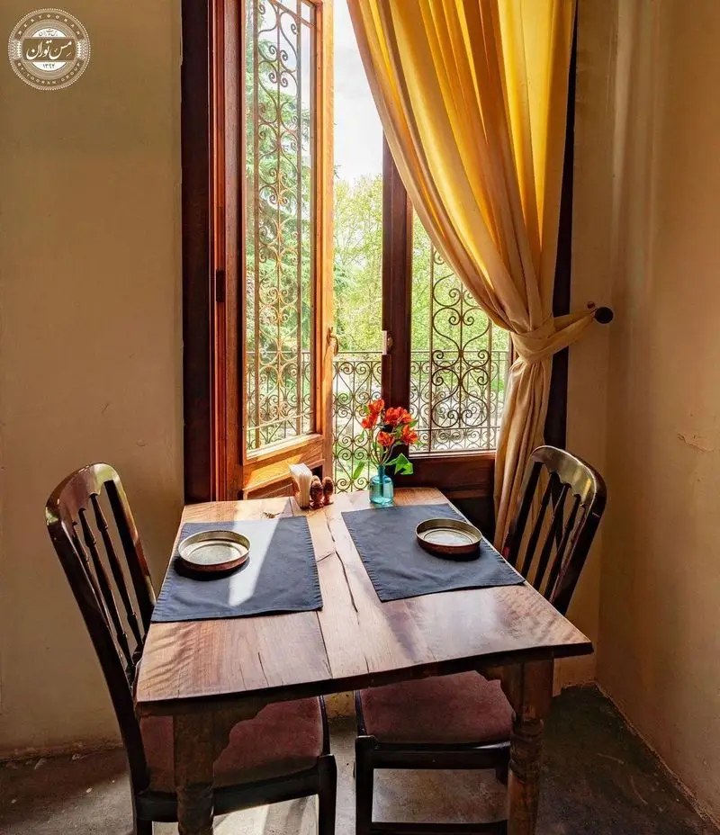 نمایی داخلی از رستوران سنتی مس توران، میز ناهارخوری دو نفره کنار پنجره‌ای با نرده‌کاری زیبا، پرده‌های زرد و گلدان گل‌های نارنجی.