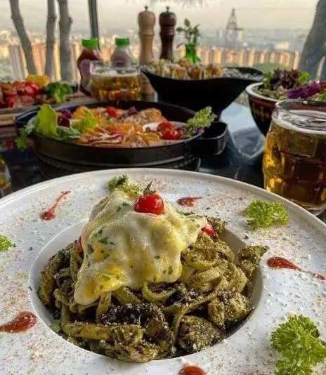 میز غذای کامل شامل پاستای پِستو با پنیر، سالاد و نوشیدنی در رستوران ایتالیایی شله با منظره شهری زیبا.