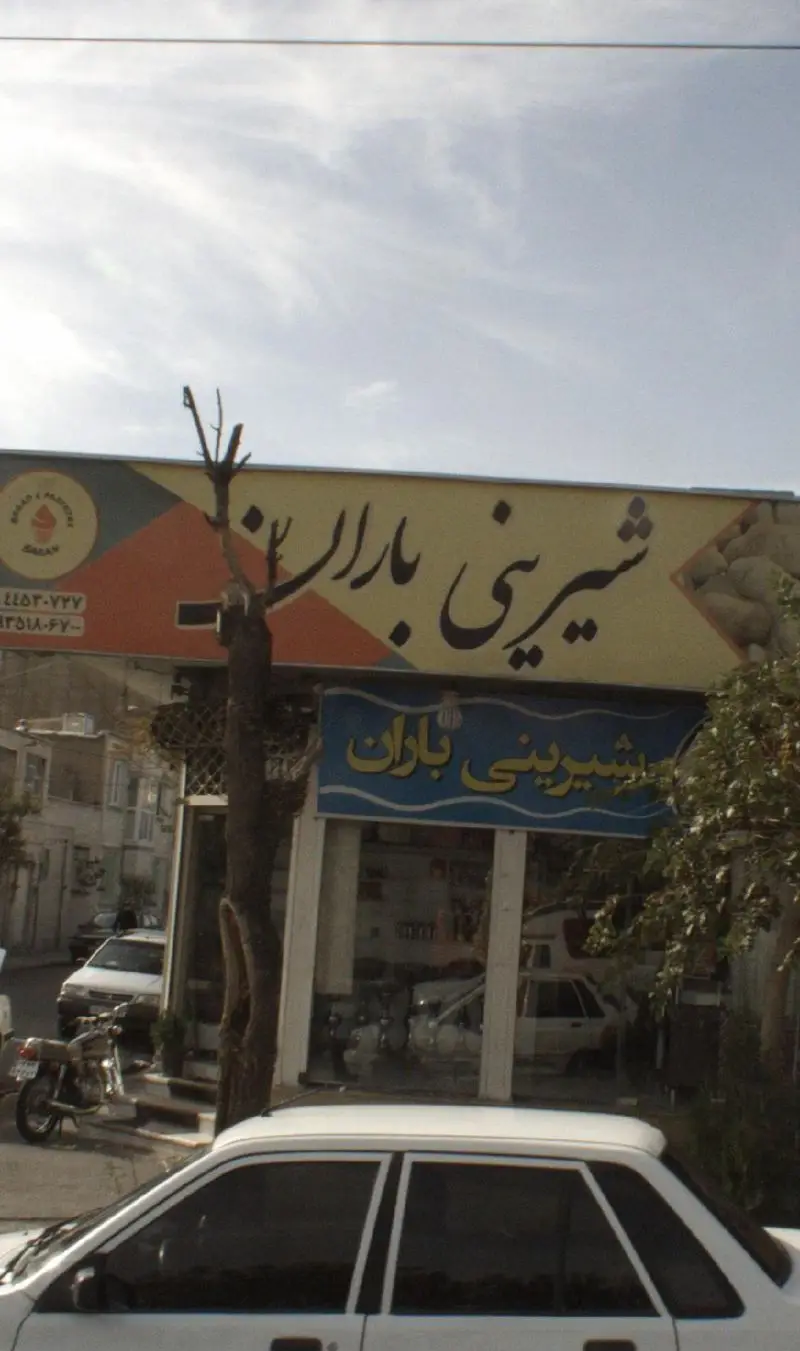 نمای بیرونی قنادی شیرینی باران با تابلوهای بزرگ زرد و آبی، شامل ماشین‌ها، موتورسیکلت و درخت در کنار خیابان شلوغ.