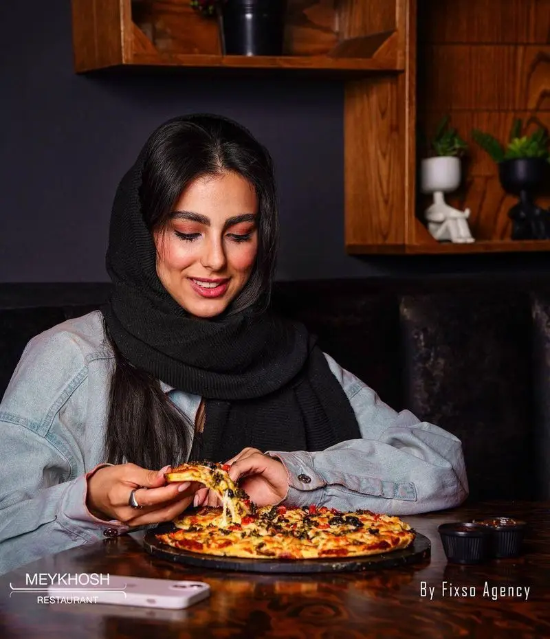 زن با حجاب در فست فود میخوش، پیتزایی خوشمزه با پنیر کشدار را از روی میز چوبی برمی‌دارد. تجربه طعم بی‌نظیر و لحظاتی شاد در رستوران.