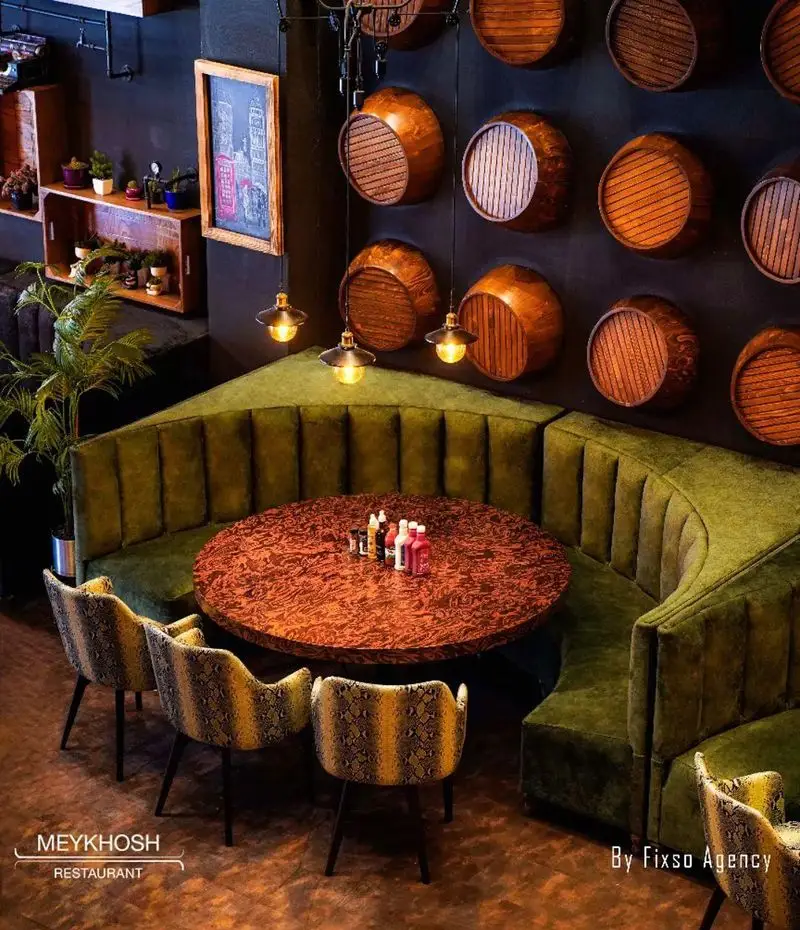 طراحی داخلی خاص فست فود میخوش: نیمکت‌های مخملی سبز، میز گرد مدرن، صندلی‌های طرح‌دار جذاب و دیوار تزئینی با بشکه‌های چوبی و نورپردازی گرم.
