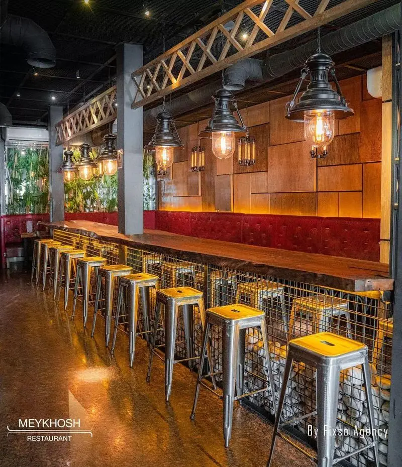 دکوراسیون داخلی چشم‌نواز فست فود میخوش با سبک صنعتی، میز بار چوبی بلند، صندلی‌های کانتر فلزی و نورپردازی گرم با چراغ‌های آویز.